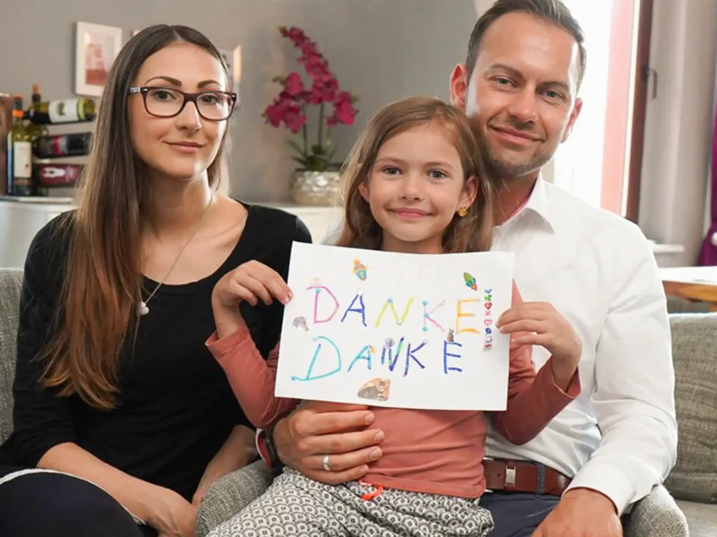 Carlotta hält ein Schild mit "Danke" darauf in die Kamera. Ihre Eltern sitzen im Hintergrund.